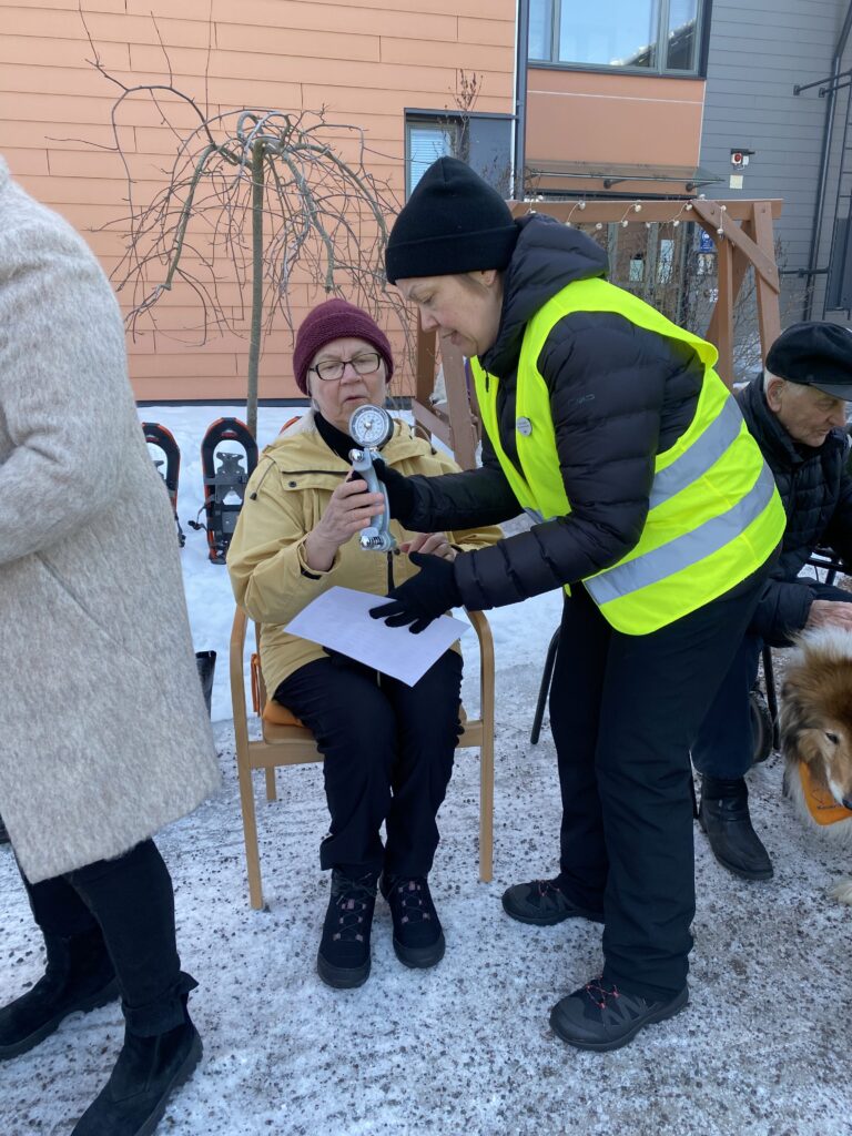 Kuvassa seniori talvitapahtumassa istumassa ja mittauttamassa käden puristusvoimaa